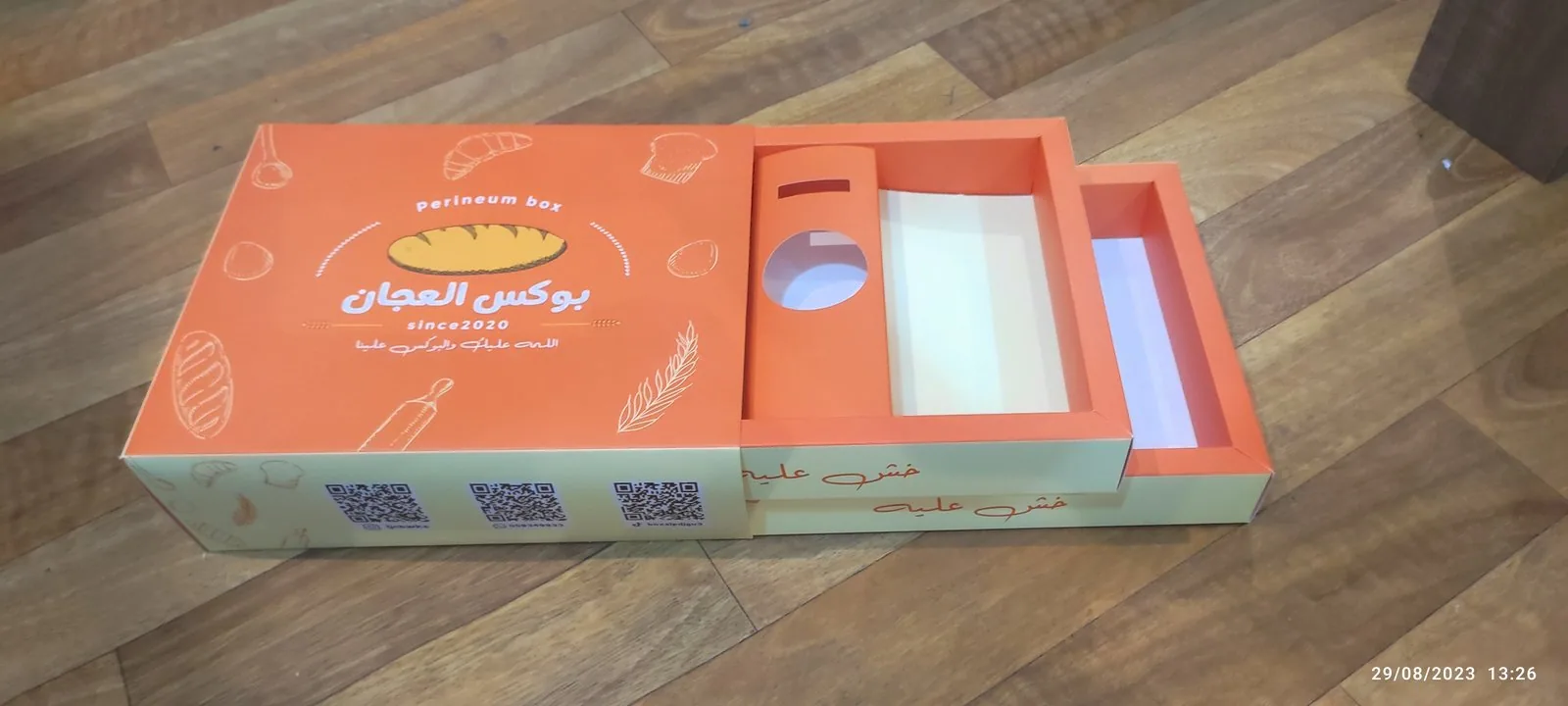 بوكس كرتوني برتقالي بنظام درج سحّاب مطبوع برسومات مخبوزات، مفتوح جزئياً ويظهر الدرج الداخلي ونافذة مفرّغة
