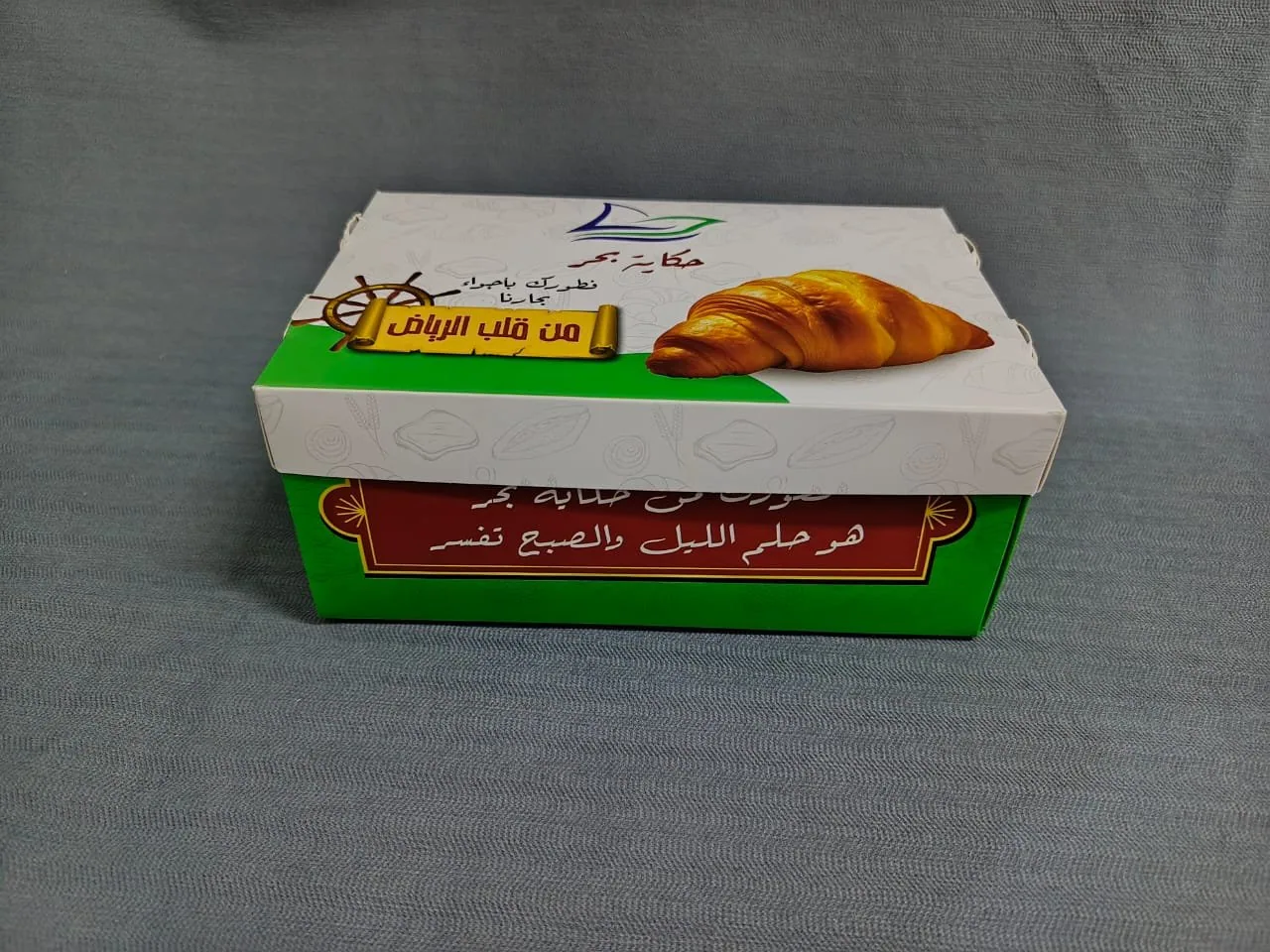بوكس كرتون صغير بغطاء منفصل مطبوع لمحل معجنات، عليه صورة كرواسون وشريط أخضر وعبارة عربية على الجانب