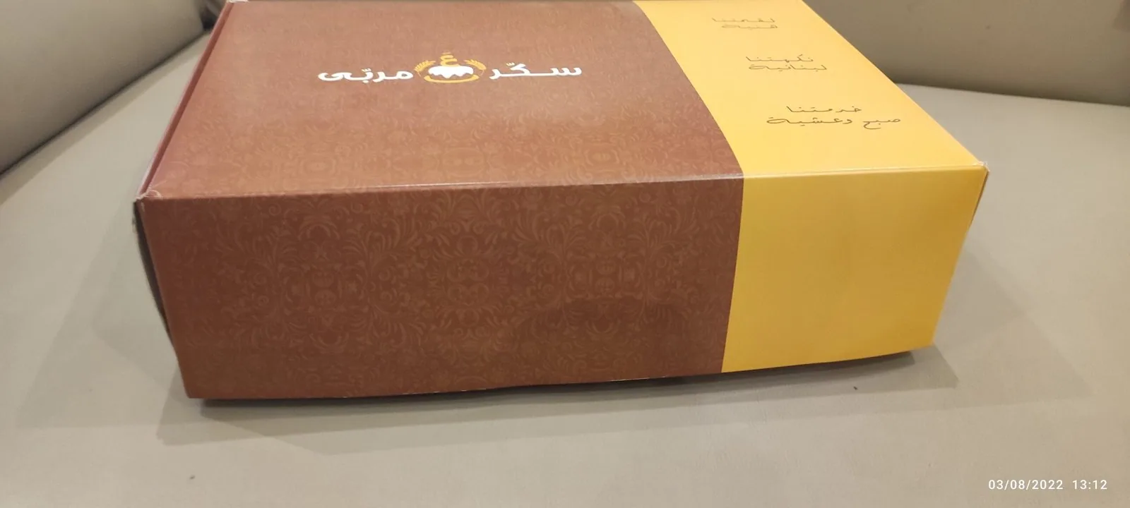بوكس كرتون مطبوع بلونين بني وأصفر مع نقش زخرفي وشعار ذهبي على الوجه العلوي، معد لتعبئة الحلويات