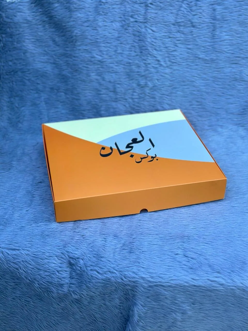 بوكس كرتوني مسطّح مطبوع بالبرتقالي والأزرق الفاتح مع اسم العلامة بخط عربي على الوجه العلوي